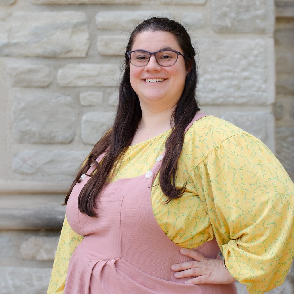 A woman with long brown hair, wearing glasses and a yellow shirt and pink jumper.