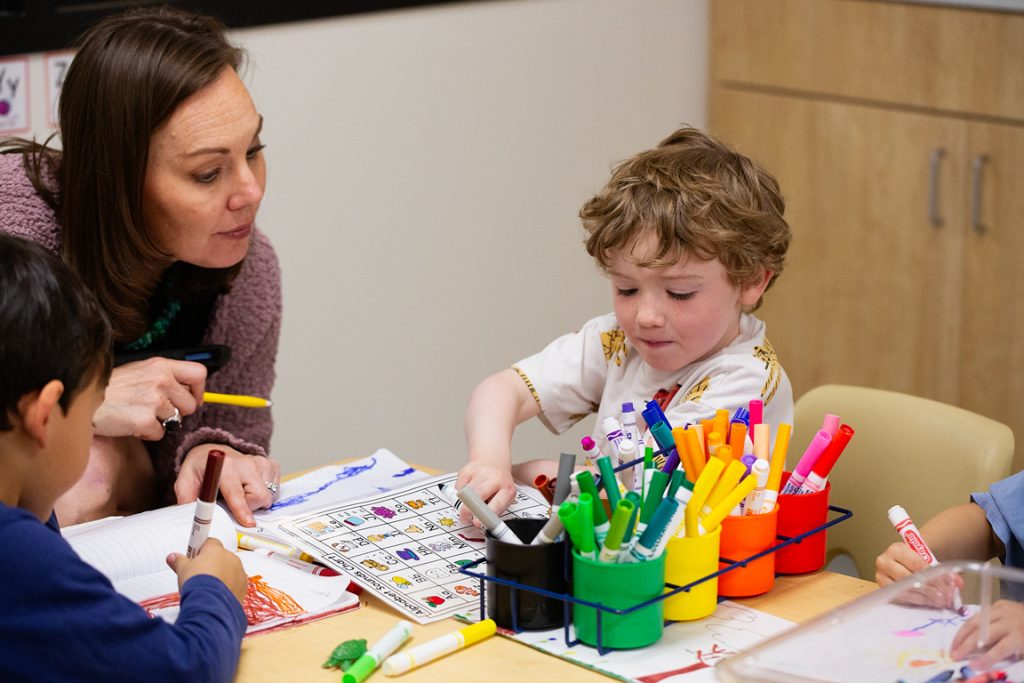 An early childhood teacher helps preschool age children at a table where they use markers to practice writing.