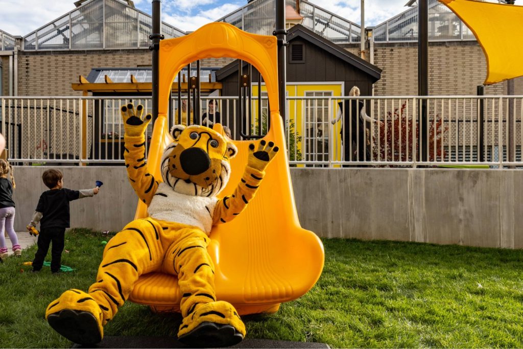 Two young children play outside at the Child Development Lab while Mizzou's mascot Truman the Tiger slides down a gold colored double slide.