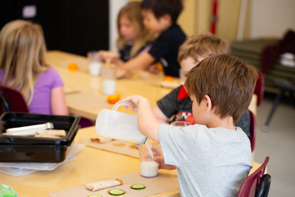 A young boy pours milk from a pitcher while seated at a lunch table with other children.