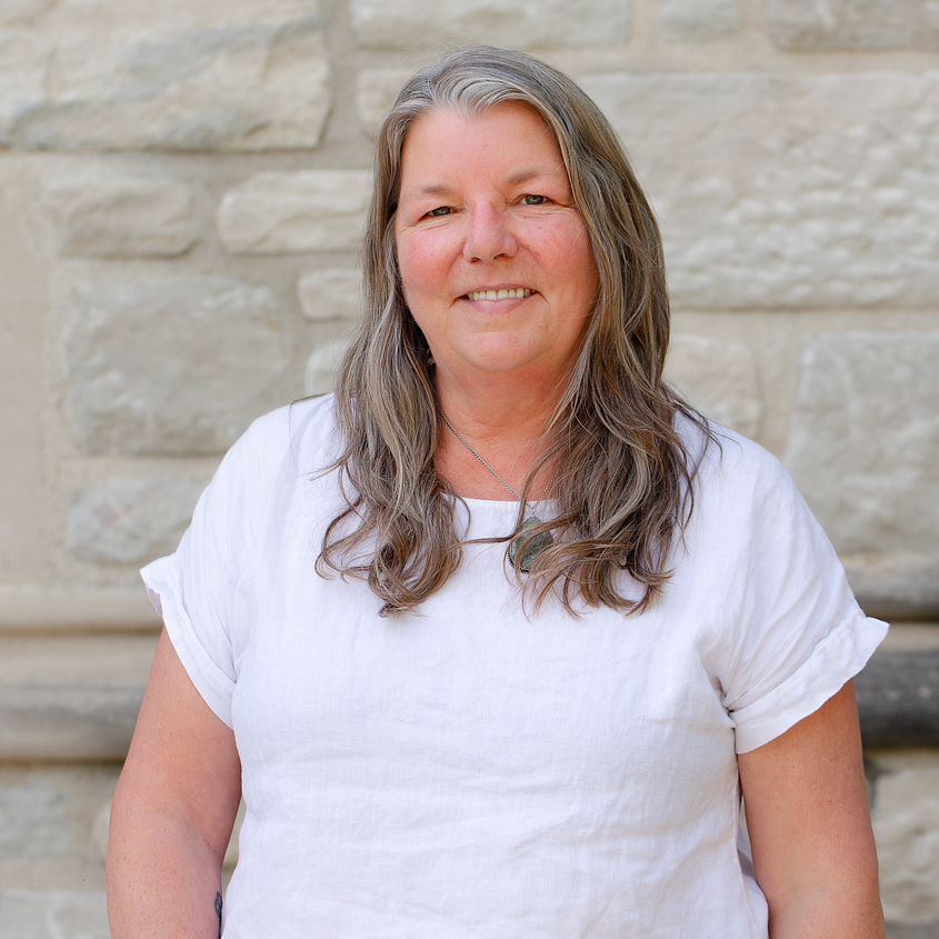 A woman with long blonde and grey hair wearing a white shirt.