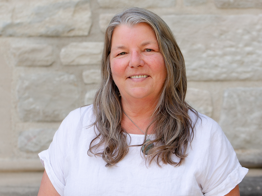 A woman with long blonde and grey hair wearing a white shirt.