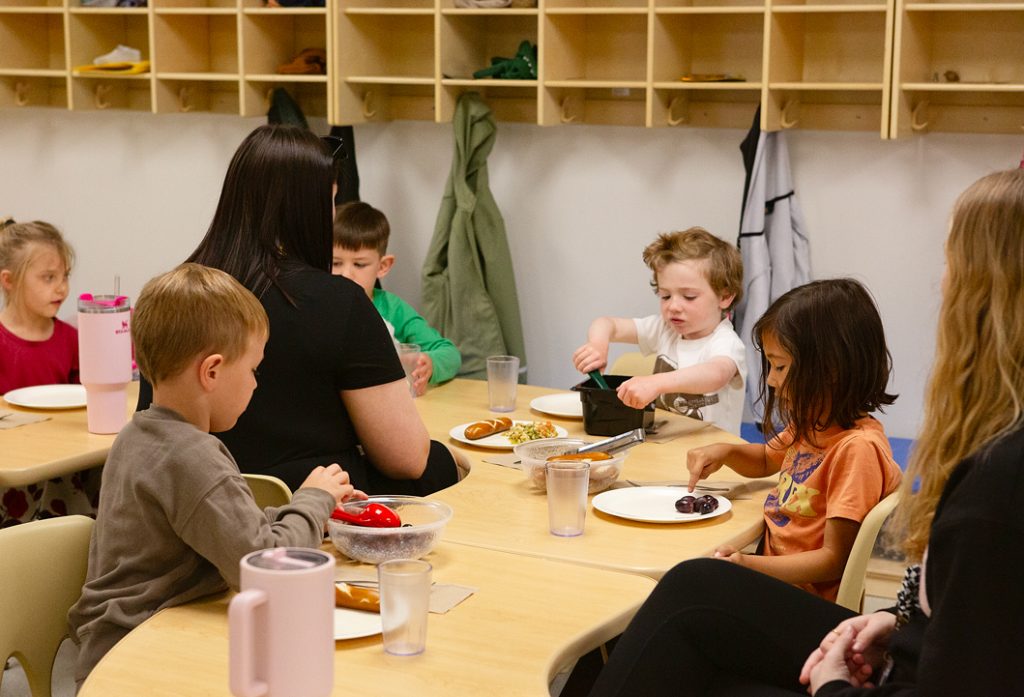 An early childhood professional sits at a table with preschool aged children as they serve lunch.